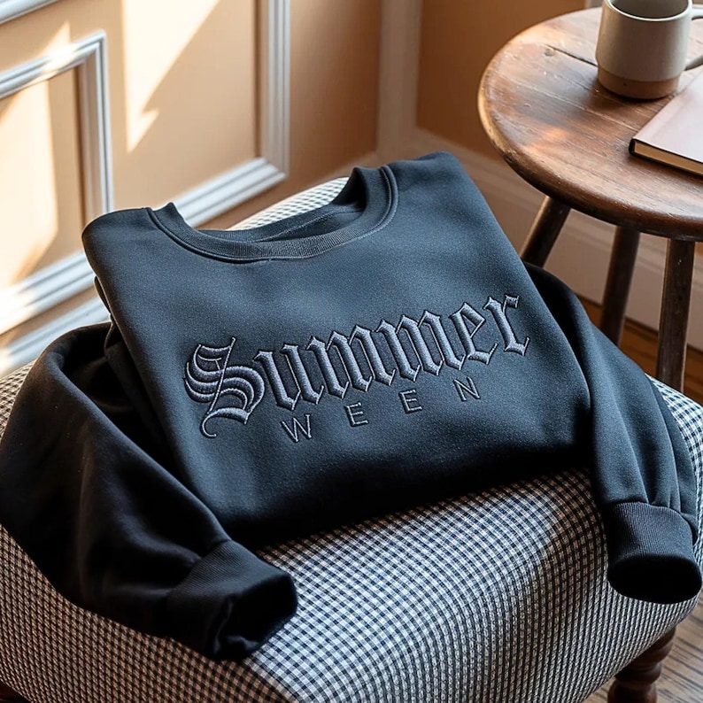 May include: A dark gray sweatshirt with the word "Summer" embroidered in a gothic font. The sweatshirt is folded on a chair with a black and white checkered pattern. A wooden side table and a mug are in the background.