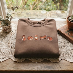 Könnte beinhalten: Ein braunes Sweatshirt mit Rundhalsausschnitt, das eine horizontale Stickerei von fünf kleinen orange-weißen Füchsen in verschiedenen Posen zeigt, darunter sitzend, zusammengerollt, stehend, springend und liegend, mittig auf der Brust.