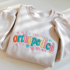 Könnte beinhalten: Beiges Sweatshirt mit dem Wort "Orthopedics" in bunten Buchstaben gestickt. Das Wort ist von gestickten Blumen und Herzen in Rosa, Gelb, Blau und Orange umgeben. Das Sweatshirt hat lange Ärmel und einen Rundhalsausschnitt.