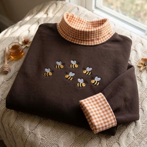 May include: A dark brown long-sleeve sweatshirt featuring a mock neck collar and sleeve cuffs in an orange and white gingham pattern. The front of the garment is embroidered with a cluster of seven small yellow and black bees.