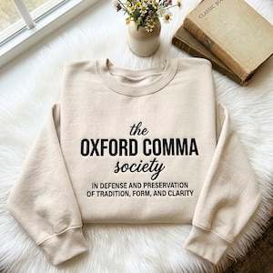 May include: A beige sweatshirt with the text "the OXFORD COMMA society" in black. Below the text is the phrase "IN DEFENSE AND PRESERVATION OF TRADITION, FORM, AND CLARITY". The sweatshirt is on a white, fluffy surface.