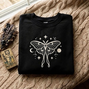May include: A black crewneck sweatshirt featuring a white line art graphic of a moth centered on the chest. The moth is surrounded by celestial elements including stars, a crescent moon, and a full moon, all rendered in a clean white outline style.