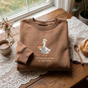 May include: A brown crewneck sweatshirt featuring an embroidered graphic of a white goose wearing a chef hat and blue apron, holding a loaf of bread and a rolling pin. Below the goose, the text reads, "sourdough goose on the loose."