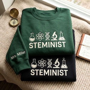 May include: A dark green sweatshirt and a black sweatshirt, both featuring white screen-printed science icons including a flask, atom, DNA strand, microscope, and beaker above the text STEMINIST. The green sleeve displays the text Mrs. Miller.