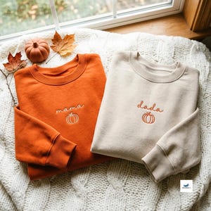 Könnte beinhalten: Zwei Rundhals-Sweatshirts, eines in gebranntem Orange und eines in Creme, jeweils mit gesticktem Text und einem Kürbis-Symbol. Das orangefarbene Sweatshirt zeigt das Wort mama, während das cremefarbene Sweatshirt das Wort dada in Schreibschrift zeigt.
