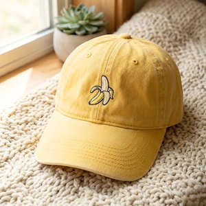 Könnte beinhalten: Senfgelbe Baseballkappe mit gebogenem Schirm. Die Kappe hat ein gesticktes Bananenmotiv. Der Hut ist aus weichem Material und liegt auf einer strukturierten Oberfläche.