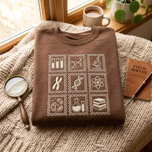 May include: A brown crewneck sweatshirt featuring a grid of nine white line-art icons resembling postage stamps. The icons depict scientific elements including test tubes, a molecular structure, a microscope, chromosomes, DNA, an atom, bacteria, a flask, and a stack of books.