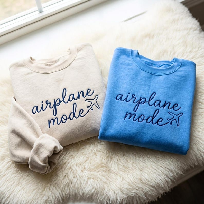 Puede incluir: Dos sudaderas de cuello redondo, una beige y otra azul claro, bordadas con las palabras "airplane mode" y un peque&ntilde;o gr&aacute;fico de avi&oacute;n azul marino. Se exhiben sobre una superficie blanca y esponjosa.