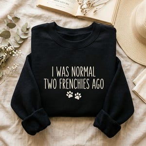 K&ouml;nnte beinhalten: Schwarzes Sweatshirt mit dem wei&szlig;en Text "I WAS NORMAL TWO FRENCHIES AGO" und zwei wei&szlig;en Pfotenabdr&uuml;cken. Das Kleidungsst&uuml;ck wird auf einer neutralen Oberfl&auml;che pr&auml;sentiert, mit einem Buch und einem Hut im Hintergrund.