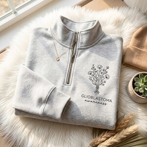 Könnte beinhalten: Hellgraues Sweatshirt mit Viertelreißverschluss, schwarzem Blumendesign und den Worten "GLIOBLASTOMA awareness". Eine goldene Halskette und eine beige Mütze sind ebenfalls sichtbar. Das Sweatshirt ist auf einer weißen, flauschigen Oberfläche gefaltet.