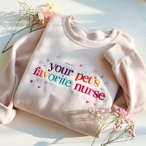 Könnte beinhalten: Hellrosa Sweatshirt mit der gestickten Aufschrift "your pet's favorite nurse" in bunten Farben. Das Design umfasst kleine Herzen, Kreuze und Sterne. Das Sweatshirt hat einen Rundhalsausschnitt und lange Ärmel.