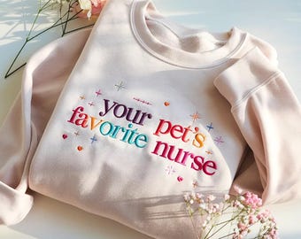May include: A light pink sweatshirt with the embroidered text "your pet's favorite nurse" in colorful thread. The design includes small hearts, crosses, and stars. The sweatshirt has a crew neck and long sleeves.