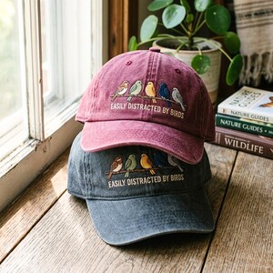 Puede incluir: Dos gorras de béisbol, una burdeos y otra gris, apiladas. Cada una presenta un diseño bordado de pájaros coloridos en una rama, con el texto "EASILY DISTRACTED BY BIRDS". Las gorras están sobre una superficie de madera.