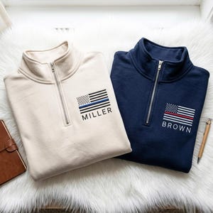 May include: Two quarter-zip pullovers, one cream and one navy blue. Each has a flag design with a coloured line and a name. The cream pullover has a blue line and the name "MILLER." The navy blue pullover has a red line and the name "BROWN."