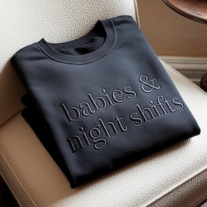 Peut inclure: Un sweat-shirt noir avec le texte brodé "babies & night shifts". Le sweat-shirt à col rond est plié et posé sur une chaise rembourrée de couleur crème. Le texte est dans une nuance plus claire de noir.
