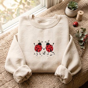 Op de afbeelding: Een beige sweater met ronde hals en een geborduurd ontwerp van twee rode lieveheersbeestjes met zwarte stippen die elkaars hand vasthouden. De lieveheersbeestjes staan in het midden op de borst, afgebeeld met dunne zwarte lijnen voor ledematen en antennes, boven klein zwart getekend gras en een piepklein bloemetje.