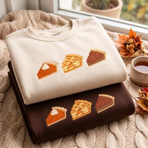 May include: A cream-coloured crewneck sweatshirt stacked on top of a dark brown crewneck sweatshirt. Both garments feature identical embroidered designs of three pie slices: a pumpkin slice with whipped cream, an apple pie slice, and a pecan pie slice.
