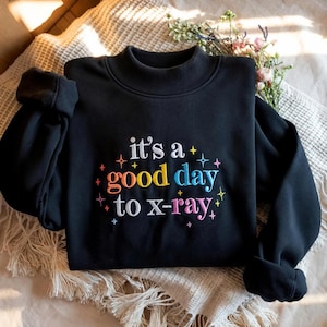 Op de afbeelding: Een zwart sweatshirt met lange mouwen en een opstaande kraag. De voorkant is voorzien van geborduurde tekst "it's a good day to x-ray" in veelkleurige letters, omringd door kleine, kleurrijke geborduurde sterretjes in roze, blauw en geel.