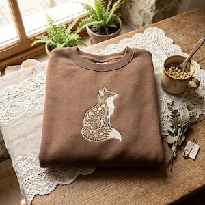 Peut inclure: Un sweat-shirt marron à col rond orné d'une broderie centrale représentant un renard assis. Le corps et la queue du renard sont remplis de motifs floraux et botaniques complexes en blanc et doré, incluant des feuilles et des petites fleurs.