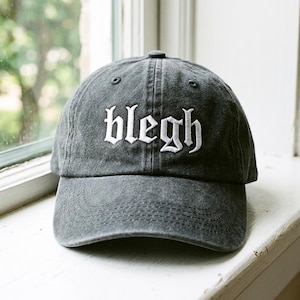 May include: A gray baseball cap with the word "blegh" embroidered in white gothic lettering. The cap is made of a soft, textured fabric and has a curved brim. The cap is sitting on a white windowsill.
