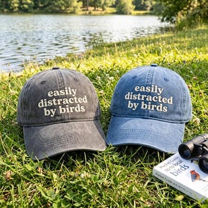 Puede incluir: Una gorra de béisbol gris carbón y otra azul denim, ambas con el texto blanco bordado "easily distracted by birds" en el panel frontal. Las gorras tienen una visera curva y una corona estructurada, expuestas junto a unos prismáticos y un libro.
