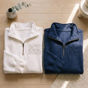 Puede incluir: Dos sudaderas con cremallera de cuarto de cierre, una blanca y otra azul marino, con "PCU NURSE" bordado en el pecho. Ambas sudaderas tienen cuello y pu&ntilde;os acanalados, y una cremallera color lat&oacute;n. Las sudaderas se exhiben sobre una superficie de madera clara.
