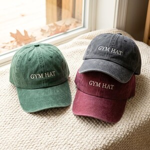 May include: Three baseball caps in shades of green, gray, and burgundy. Each cap has the text "GYM HAT" embroidered in white. The caps are arranged on a cream-colored, textured surface near a window.