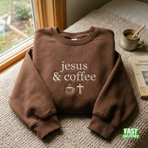 Può includere: Una felpa marrone a girocollo con il testo ricamato "jesus & coffee" in color crema chiaro. Sotto il testo, è presente un piccolo ricamo raffigurante una tazza di caffè fumante accanto al simbolo di una semplice croce cristiana.