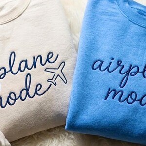 Puede incluir: Dos sudaderas de cuello redondo, una beige y otra azul claro, bordadas con las palabras "airplane mode" y un peque&ntilde;o gr&aacute;fico de avi&oacute;n. Las sudaderas son suaves e informales, perfectas para viajar.