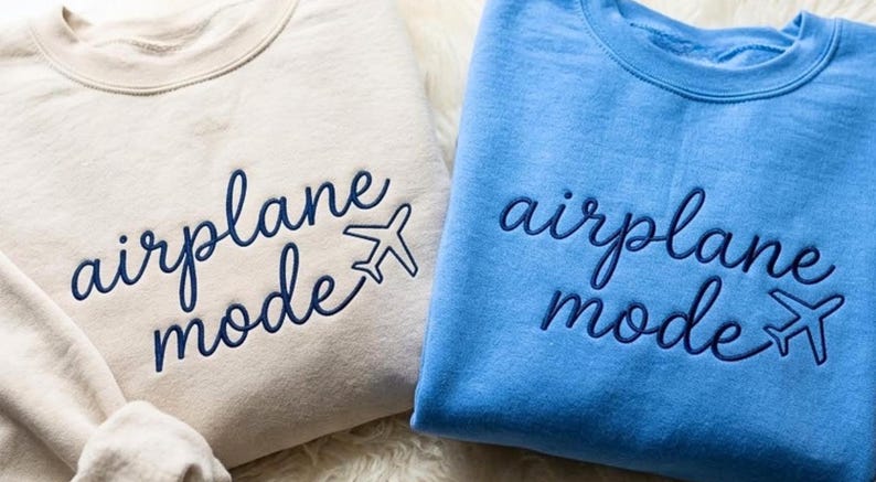 Puede incluir: Dos sudaderas de cuello redondo, una beige y otra azul claro, bordadas con las palabras "airplane mode" y un peque&ntilde;o gr&aacute;fico de avi&oacute;n. Las sudaderas son suaves e informales, perfectas para viajar.
