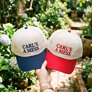 Puede incluir: Dos gorras de béisbol con el texto "CARL'S A MESS". Una gorra tiene una visera azul marino y la otra una visera roja. Ambas gorras tienen una corona beige y un botón en la parte superior.