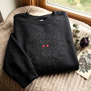 May include: A black crewneck sweatshirt featuring an embroidered design of a moth-like creature with two bright red circular eyes. The text MOTHMAN is stitched in an arch above the creature, surrounded by small embroidered stars, clouds, and moons.