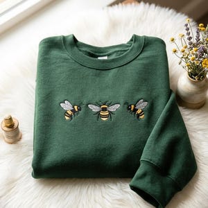 Pode incluir: Camisola verde escuro com três abelhas bordadas no peito. As abelhas são amarelas, pretas e brancas, com asas detalhadas. A camisola está dobrada sobre uma superfície branca e fofa.