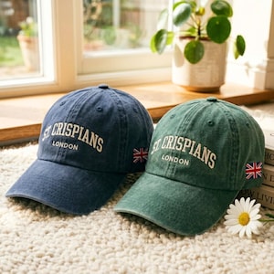 Puede incluir: Dos gorras de béisbol, una azul marino y otra verde, están una al lado de la otra. Ambas gorras tienen el texto "ST CRISPIANS LONDON" en blanco, con una pequeña bandera de la Union Jack. Están sobre una superficie suave y texturizada.