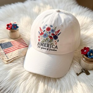 May include: White baseball cap with embroidered floral design in red, blue, and green. The cap features the word "AMERICA" and the text "250 years of freedom". The cap is a patriotic accessory.