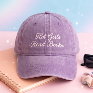May include: A lavender baseball cap with the embroidered text "Hot Girls Read Books." The cap is made of a soft, washed fabric and has a curved brim. The hat is sitting on a pink notebook.