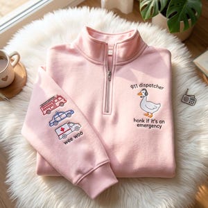 May include: Pale pink quarter-zip sweatshirt with embroidered emergency vehicle graphics on the left sleeve. The right chest features a goose wearing a headset with the text "911 dispatcher, honk if it's an emergency."