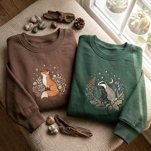 Könnte beinhalten: Ein braunes Sweatshirt mit einem gestickten Fuchs, umgeben von Blumen und Sternen, und ein grünes Sweatshirt mit einem gestickten Dachs, umgeben von Farnen, Blumen und einer Mondsichel. Beide Kleidungsstücke haben einen Rundhalsausschnitt.