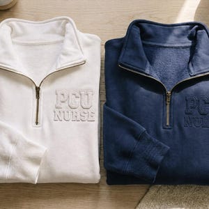 Puede incluir: Dos sudaderas con cremallera de un cuarto, una blanca y otra azul marino, con el texto bordado "PCU NURSE". Ambas sudaderas tienen cuello y pu&ntilde;os acanalados, y una cremallera color lat&oacute;n. Las sudaderas est&aacute;n hechas de un material suave y c&oacute;modo.
