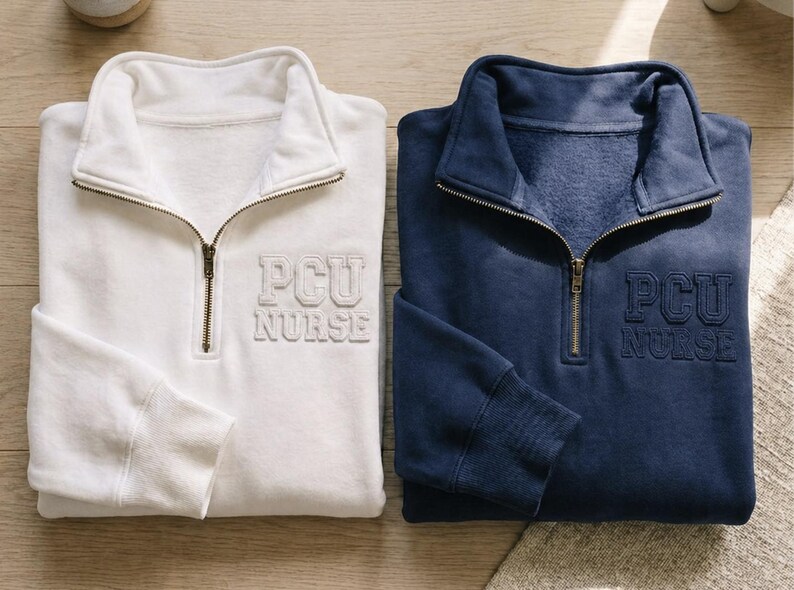 Puede incluir: Dos sudaderas con cremallera de un cuarto, una blanca y otra azul marino, con el texto bordado "PCU NURSE". Ambas sudaderas tienen cuello y pu&ntilde;os acanalados, y una cremallera color lat&oacute;n. Las sudaderas est&aacute;n hechas de un material suave y c&oacute;modo.