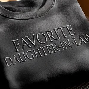May include: A black sweatshirt with the embroidered text "FAVORITE DAUGHTER-IN-LAW" in a light grey color. The sweatshirt has a crew neck and a soft, textured appearance. The image is a close-up shot.