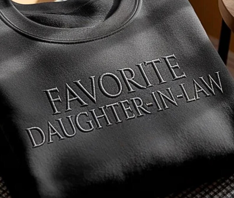 May include: A black sweatshirt with the embroidered text "FAVORITE DAUGHTER-IN-LAW" in a light grey color. The sweatshirt has a crew neck and a soft, textured appearance. The image is a close-up shot.