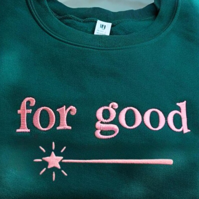K&ouml;nnte beinhalten: Ein dunkelgr&uuml;nes Sweatshirt mit dem gestickten Schriftzug "for good" in Rosa, begleitet von einem rosa Stern und Zauberstab-Design. Das Sweatshirt hat einen Rundhalsausschnitt und ein kleines Etikett am Ausschnitt.