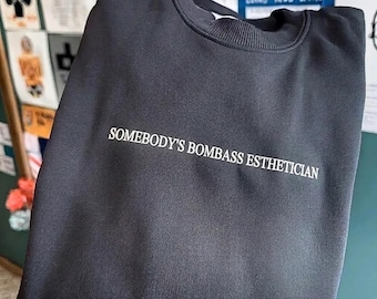 May include: Black crewneck sweatshirt with the text "SOMEBODY'S BOMBASS ESTHETICIAN" in white. The sweatshirt has a ribbed collar and a soft, casual appearance. The garment is likely made of a comfortable fabric, suitable for everyday wear.