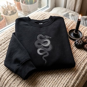 Può includere: Una felpa nera a girocollo con un dettagliato ricamo bianco raffigurante un serpente attorcigliato al centro. Il disegno mette in risalto le squame del serpente e la lingua biforcuta sulla parte anteriore del capo a maniche lunghe con polsini.