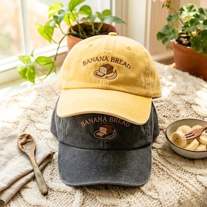 May include: Two baseball caps, one yellow and one dark gray, stacked on top of each other. Both caps feature the embroidered text "BANANA BREAD" above an illustration of a banana and a slice of bread. The caps are on a cream-colored, textured surface.