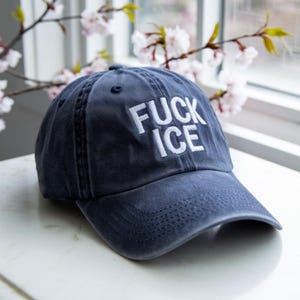 Könnte beinhalten: Marineblaue Baseballkappe mit den Worten "FUCK ICE" in Weiß gestickt. Die Kappe hat einen gebogenen Schirm und ein klassisches Sechs-Panel-Design. Der Hut liegt auf einer weißen Oberfläche.
