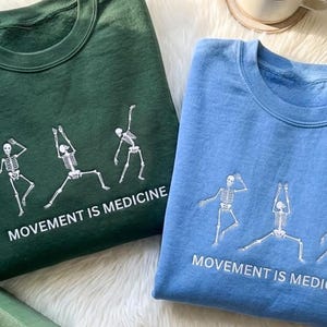May include: Two sweatshirts, one teal and one light blue, are displayed. Each features a white skeleton graphic in various yoga poses, with the text "MOVEMENT IS MEDICINE" below. The sweatshirts are folded, showcasing the design.