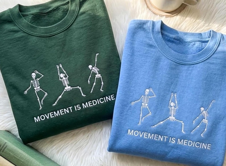 May include: Two sweatshirts, one teal and one light blue, are displayed. Each features a white skeleton graphic in various yoga poses, with the text "MOVEMENT IS MEDICINE" below. The sweatshirts are folded, showcasing the design.