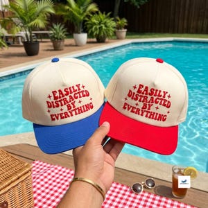 May include: Two baseball caps with off-white crowns and red embroidered text that reads EASILY DISTRACTED BY EVERYTHING with small star accents. One cap features a solid blue brim, while the other cap features a matching solid red brim.